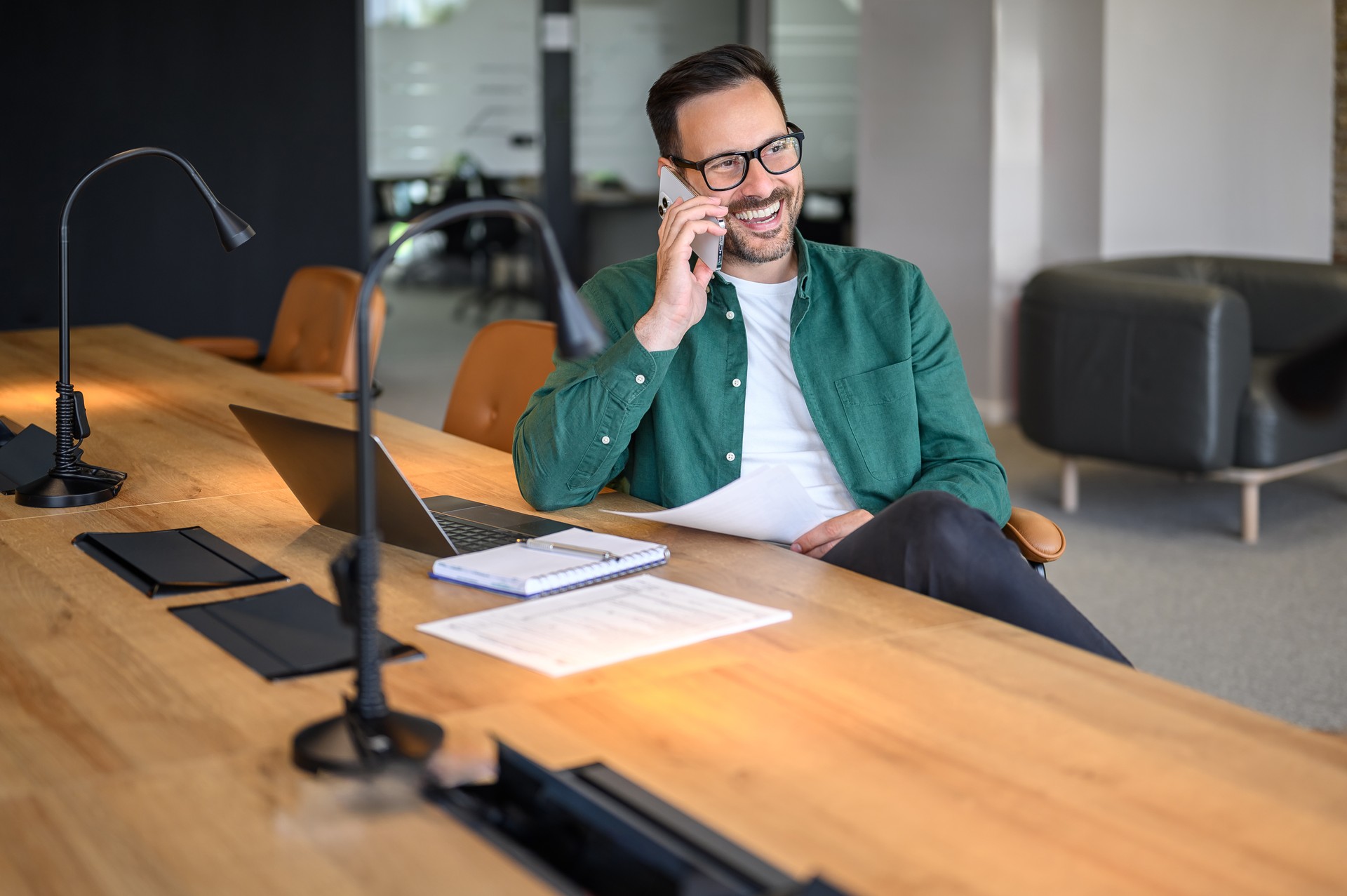 Glücklicher erwachsener Geschäftsmann, der per Telefonanruf diskutiert und wegschaut, während er am Schreibtisch im Büro sitzt